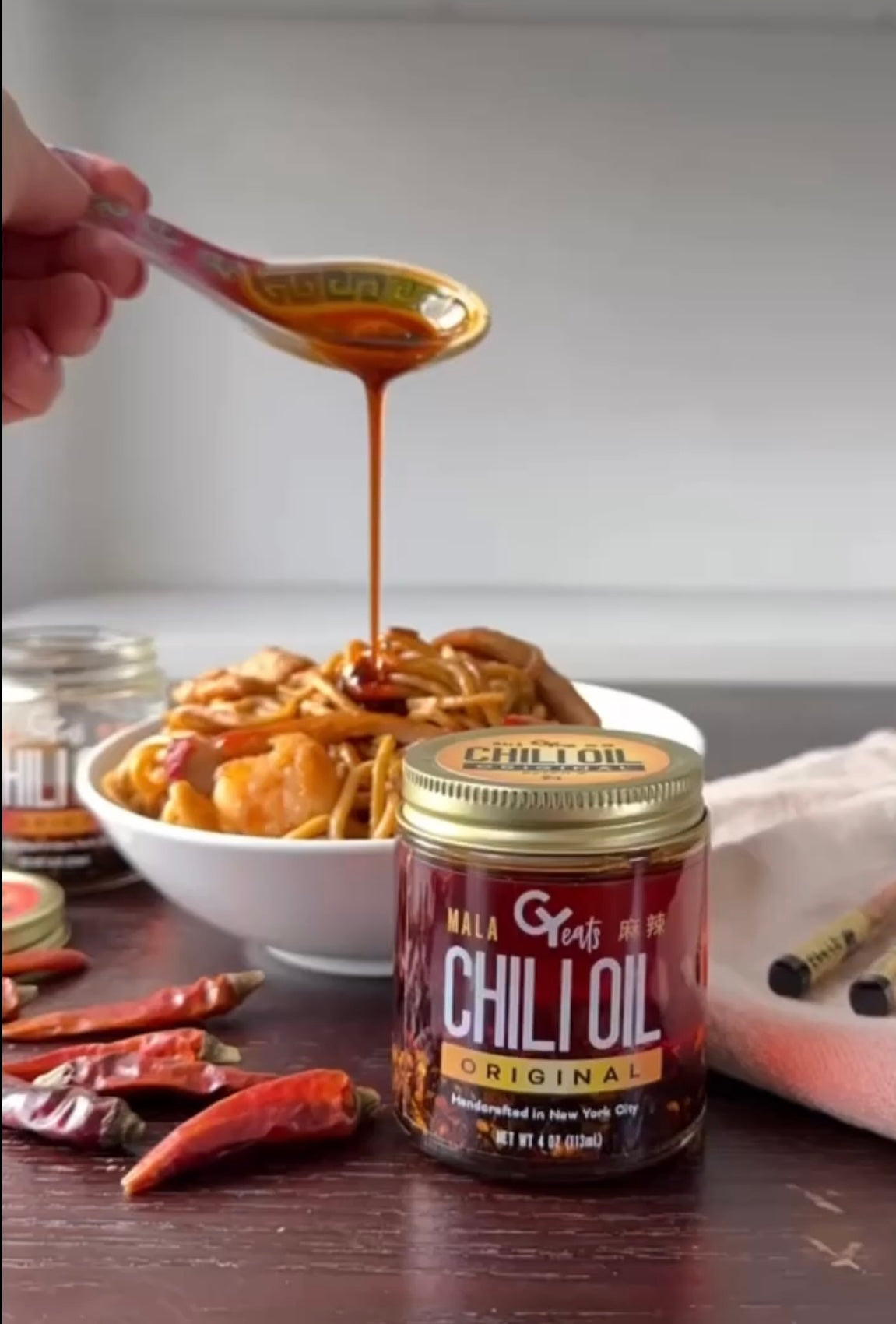 Pouring chili oil from a spoon into noodles, with Original chili oil jar and gold lid
