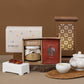 A gift set arrangement featuring a SEOGA-branded box with a jar and red packet, alongside a white SEOGA box, a lit wooden lantern, a white teapot, a cup of tea, and small amber-colored stones on a warm brown background.