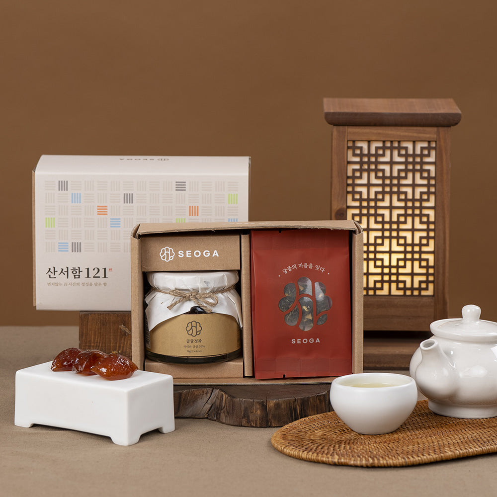 A gift set arrangement featuring a SEOGA-branded box with a jar and red packet, alongside a white SEOGA box, a lit wooden lantern, a white teapot, a cup of tea, and small amber-colored stones on a warm brown background.