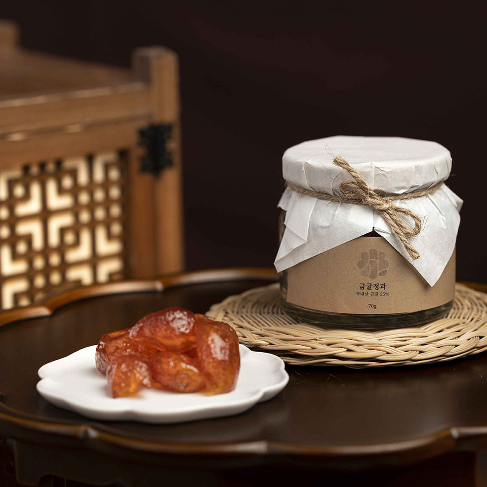 A glass jar of honey with a white paper cover tied with twine sits on a woven mat beside a small plate holding a piece of candied fruit, set on a dark wooden table with a decorative wooden chair in the background.
