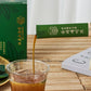 Gombo baechu syrup being poured into a glass cup, with green boxes nearby.