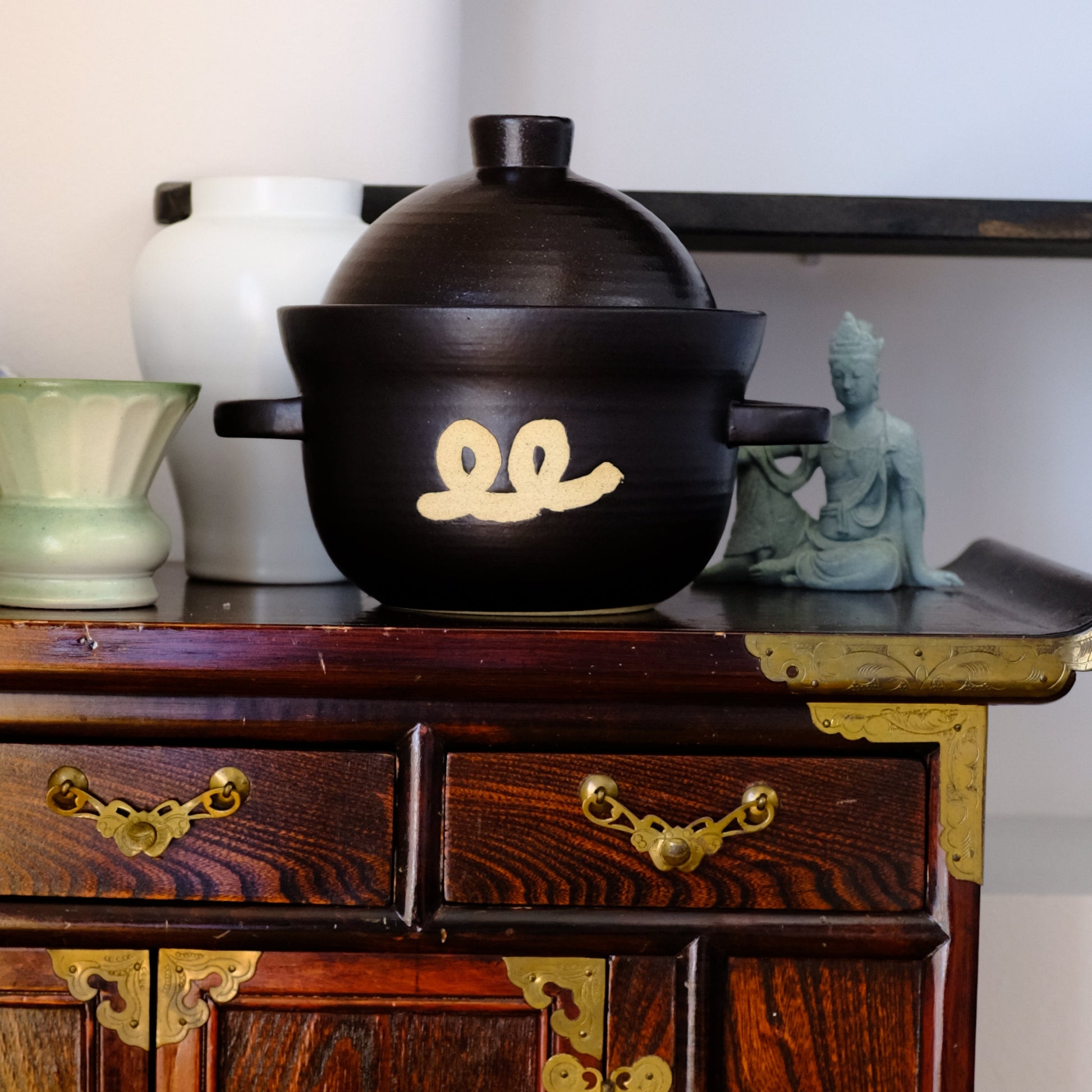 Black ceramic rice pot with lid, gold swirl emblem and side handles.