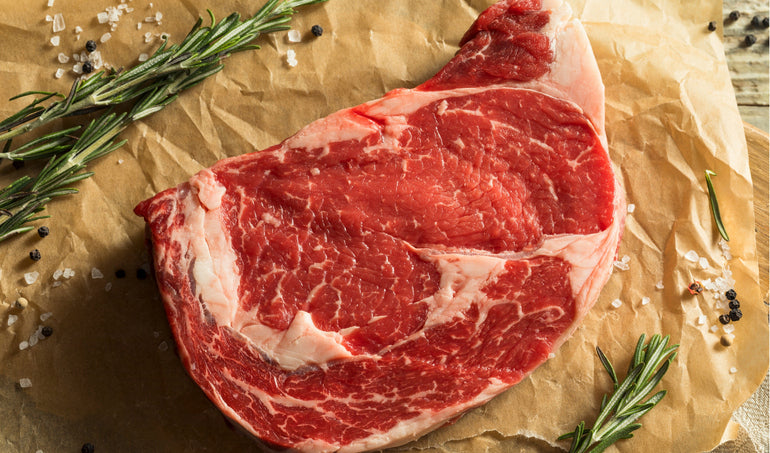 Raw ribeye steak on parchment with herbs and peppercorns