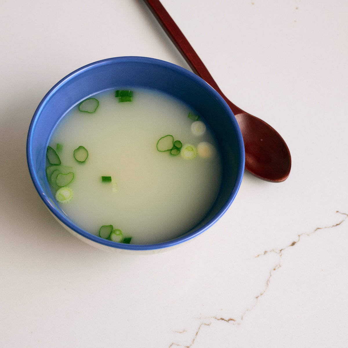 Blue bowl of pale bone broth with sliced green onions and spoon