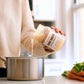 Jar of bone broth being poured into a stainless pot with herbs nearby
