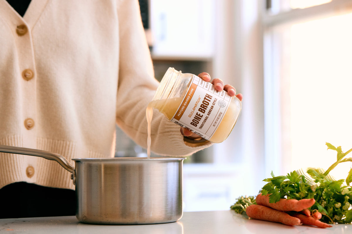 Jar of bone broth being poured into a stainless pot with herbs nearby