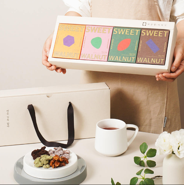 A person in a beige apron holds a white gift box labeled 'SWEET WALNUT' with four colorful flavor panels. A matching white gift bag sits on the table, alongside a white mug, a small dish of assorted nuts, and white flowers with green leaves.