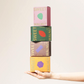Five colorful Sweet Walnut snack boxes are stacked vertically on a person's hand against a plain light beige background. The boxes are pink, green, brown, and yellow, each featuring a different geometric shape in the center and the words 'SWEET WALNUT' printed vertically on the sides.