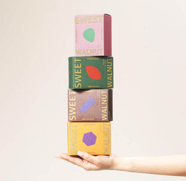 Five colorful Sweet Walnut snack boxes are stacked vertically on a person's hand against a plain light beige background. The boxes are pink, green, brown, and yellow, each featuring a different geometric shape in the center and the words 'SWEET WALNUT' printed vertically on the sides.