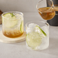 Two clear rocks glasses filled with a clear bubbly cocktail and lime slices, one resting on a round coaster and one being poured into from a small glass pitcher held by a hand. A bottle and other bar items are blurred in the background on a light gray surface.