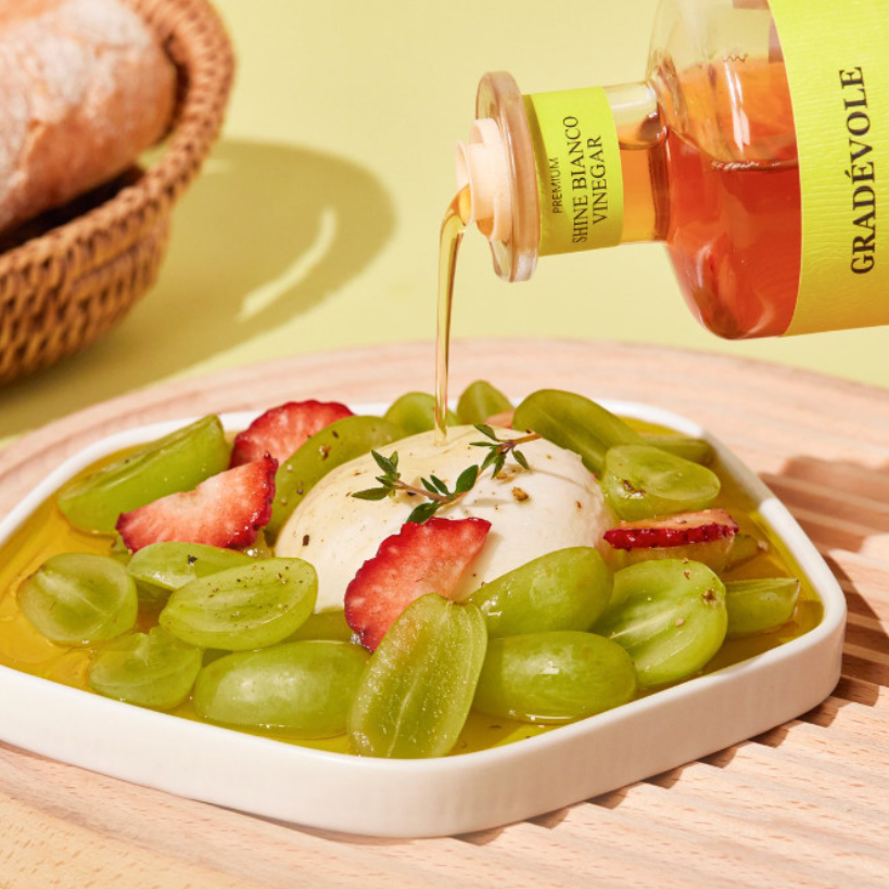 A close-up of a plated dessert topped with green grapes, sliced strawberries, and a dollop of cream, with golden syrup being poured from a bottle labeled Gradévôle. The scene is styled on a wooden board with a soft yellow-green background and a blurred loaf of bread in a basket.