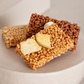 Three square rice crispy snack bars with different toppings, including dried fruit slices and a white candy piece, arranged on a light beige ceramic plate.