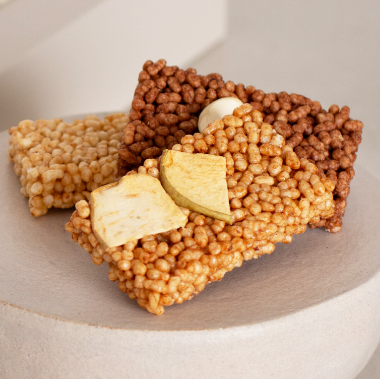 Three square rice crispy snack bars with different toppings, including dried fruit slices and a white candy piece, arranged on a light beige ceramic plate.