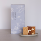 A rectangular light gray box with white botanical line art stands behind a small ceramic dish holding two square blocks of textured rice cake, one light beige and one medium brown, on a neutral gray surface.