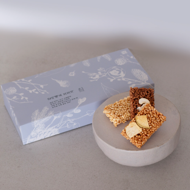 A light blue rectangular box with white botanical line art sits behind two square rice crispy snack bars on a round gray pedestal. The snack bars are topped with sesame seeds, cashews, and dried fruit pieces.