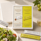 Gradévole Shude Balsamic Vinegar set displayed with its white gift box, sachet pack, and individual tubes on a light beige surface, styled with a green salad, a glass of balsamic, and green grapes.