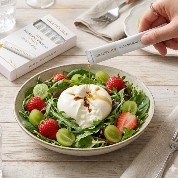 A hand pours a single dose of Gradévøle Shreve Balanced Sugar drink mix from a tube into a bowl of fresh salad with a ball of mozzarella, strawberries, and grapes. The product box is visible in the background on a light wood table.