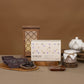 A styled product arrangement on a warm brown surface against a matching brown backdrop. The main subject is a rectangular product box with Korean text, shown alongside several dark pouches, a small jar with a white lid and string, and wooden serving pieces.