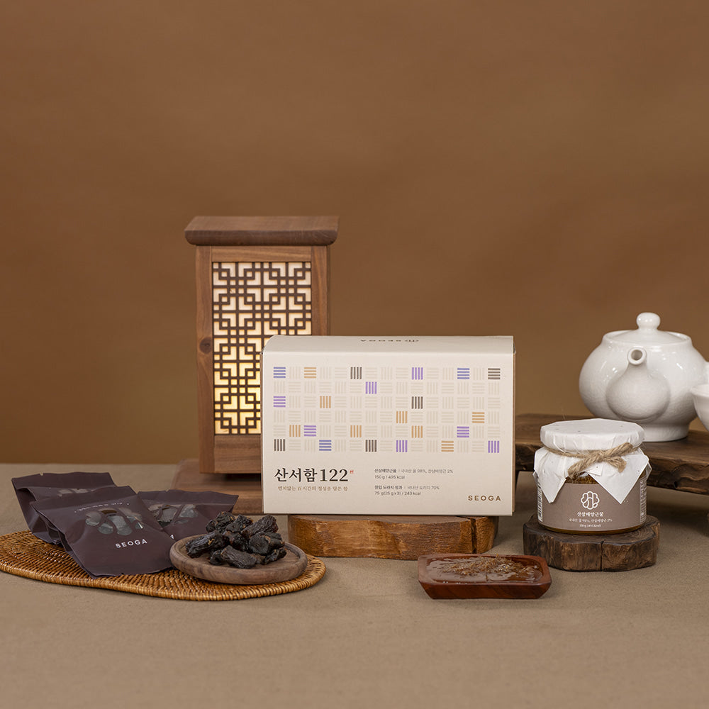 A styled product arrangement on a warm brown surface against a matching brown backdrop. The main subject is a rectangular product box with Korean text, shown alongside several dark pouches, a small jar with a white lid and string, and wooden serving pieces.